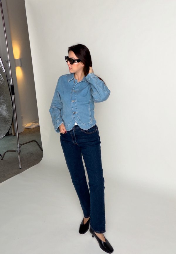 Una mujer posa en un estudio fotográfico con gafas de sol negras, una camisa vaquera azul claro, vaqueros oscuros y zapatos negros de tacón.