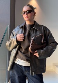 Bolsa de ombro em couro castanho com uma forma curvada, sobreposta a um suéter cinza, combinada com uma jaqueta de couro preta e jeans escuros.