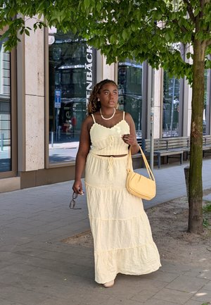 Żółta, warstwowa sukienka maxi z dopasowanym topem, regulowanymi ramiączkami i talią ze sznurkiem; w połączeniu z jas yellow torebką i okularami przeciwsłonecznymi.
