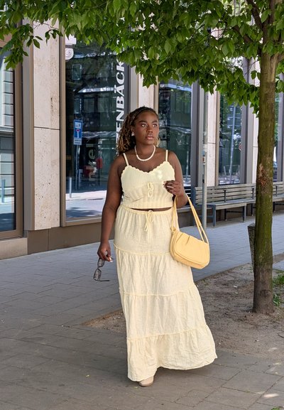 Vestido maxi amarillo de volantes con un top ajustado, tirantes ajustables y cintura con cordón; combinado con un bolso amarillo claro y gafas de sol.
