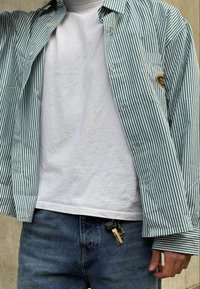 Ριγέ πουκάμισο σε πράσινο και λευκό, φορεμένο πάνω από ένα απλό λευκό μπλουζάκι, με μπλε τζιν παντελόνι. Κλειδιά ορατά προσαρτημένα στη ζώνη.