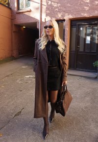 Cappotto lungo marrone sopra un maglione corto nero, abbinato a una gonna nera. Tacchi lucidi a punta, calze nere e una borsa marrone completano il look.