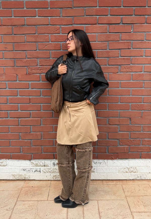 Mujer con cabello largo y oscuro que lleva gafas, una chaqueta de cuero negra, falda beige, pantalones marrones y sostiene un bolso marrón, de pie junto a una pared de ladrillos.