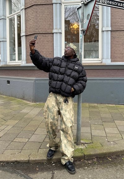 Cazadora negra tipo puffer, pantalones holgados de camuflaje estampado, zapatos negros; persona haciéndose un selfie cerca de una señal de tráfico en un entorno urbano.