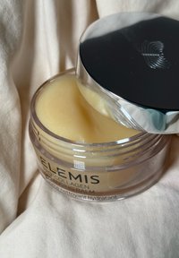 Tarro abierto de Elemis Pro-Collagen Cleansing Balm con una suave crema amarilla en su interior, reposando sobre una tela beige.