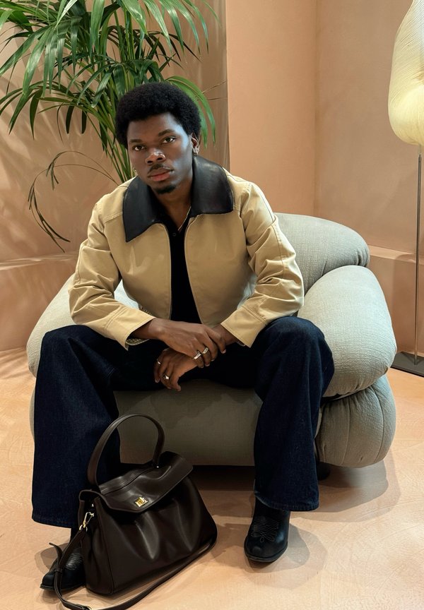 Beige Jacke mit schwarzem Lederkragen, schwarzes Poloshirt, dunkelblaue Jeans, dunkle Handtasche mit goldener Schnalle, sitzt auf einem plüschigen, hellen Sofa.