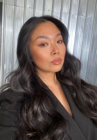 Svart blazer, bølgete svart hår, jevn hudtone, myk sminke med glossede lepper, bakgrunnen har metalliske vertikale linjer.