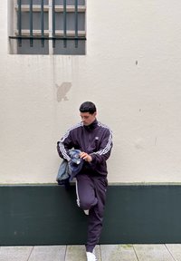 Survêtement noir avec des rayures blanches, ajustant ses vêtements, debout contre un mur de couleur claire avec une fenêtre barrée au-dessus.