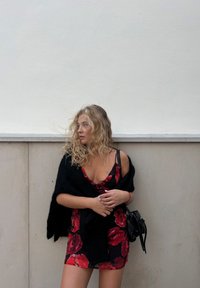 Rotes Blumenkleid mit taillierter Form und schwarzen Akzenten, kombiniert mit einem schwarzen Schal. Glattes Material mit leichtem Glanz. Kleine schwarze Tasche.