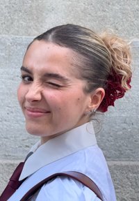 Giovane con capelli castano chiaro, mossi, raccolti in uno chignon, che indossa una camicia bianca e una cravatta color bordeaux. Accessorio per capelli rosso e orecchini d'argento.