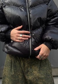 Crna skakačka jakna sa sjajnim završetkom, patentnim zatvaračem i elastikom na manžetama, u kombinaciji s pantalonama s kamuflažnim uzorkom. Ruke ukrašene srebrnim prstenjem.