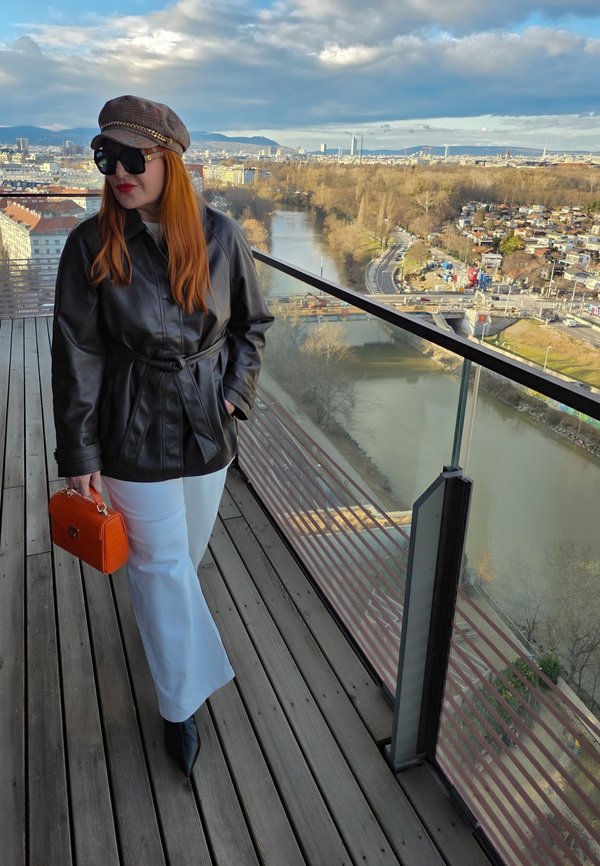 Vrouw met rood haar die een leren jas, witte broek, geruit petje, zonnebril draagt en een oranje handtas vasthoudt op een balkon met uitzicht op een rivier en stadsgezicht.