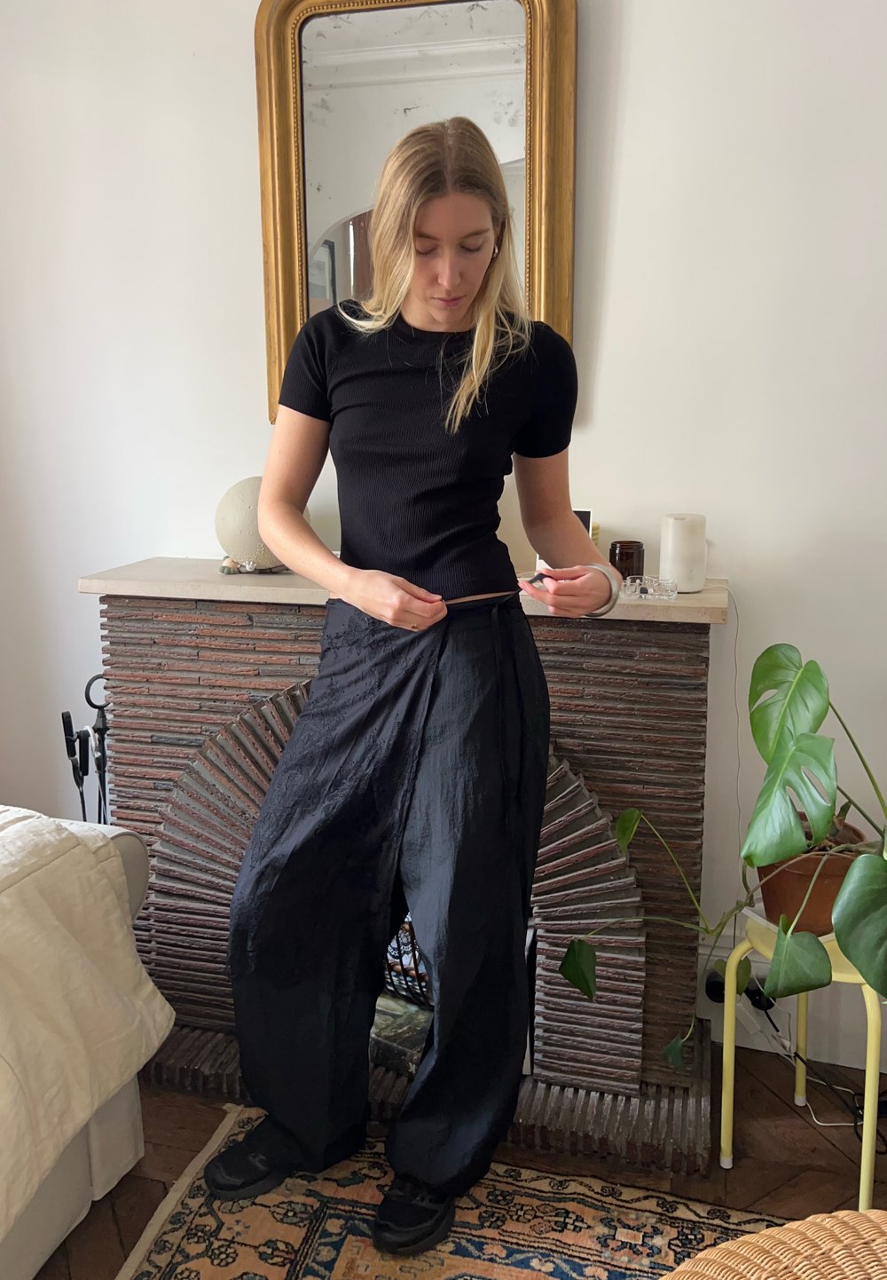 Must have mustriba lühikeste varrukatega tsärk, mis on kombineeritud kergete mustade pükstega; taustal on tekstureeritud kivikamin ja taimed.