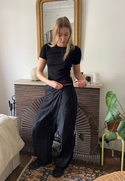 Top negro de manga corta con textura de canalé combinado con pantalones anchos negros de tejido ligero; el entorno presenta una chimenea de piedra texturizada y plantas.