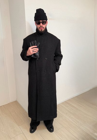 Hombre con un abrigo largo negro, gorro negro, gafas de sol y zapatos negros, sosteniendo guantes de cuero negros contra un fondo de pared blanca.