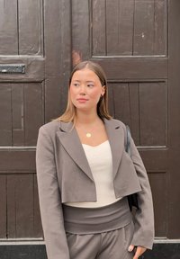 Siva skrajšana blazer jakna čez belo rebrovano majico, v kombinaciji z usklajenimi sivimi hlačami. Obleka ima subtilno teksturo in minimalen dizajn.