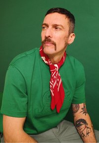 Camiseta verde com design bordado, bandana vermelha com padrão branco, mangas curtas e antebraço tatuado em fundo verde.