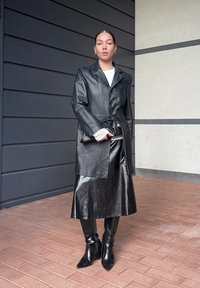 Manteau en cuir noir à coupe ajustée, associé à une jupe noire brillante et des bottes hautes jusqu'aux genoux, présenté sur un fond neutre.
