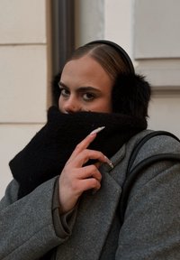 Zwarte nepbont oorkappen en een dikke zwarte sjaal worden gedragen bij een grijze wollen jas. De sjaal hangt om de nek en bedekt gedeeltelijk het gezicht.