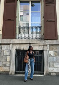 Top nero a collo alto, jeans azzurri a vita alta, borsa marrone grande, sciarpa a fantasia, scarpe con tacco quadrato; in piedi di fronte a un muro di pietra.