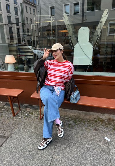 Camisa a rayas rojas y blancas, chaqueta bomber oscura, jeans azules, zapatillas multicolores, gorra beige, llavero de peluche, sentado en un banco de madera.