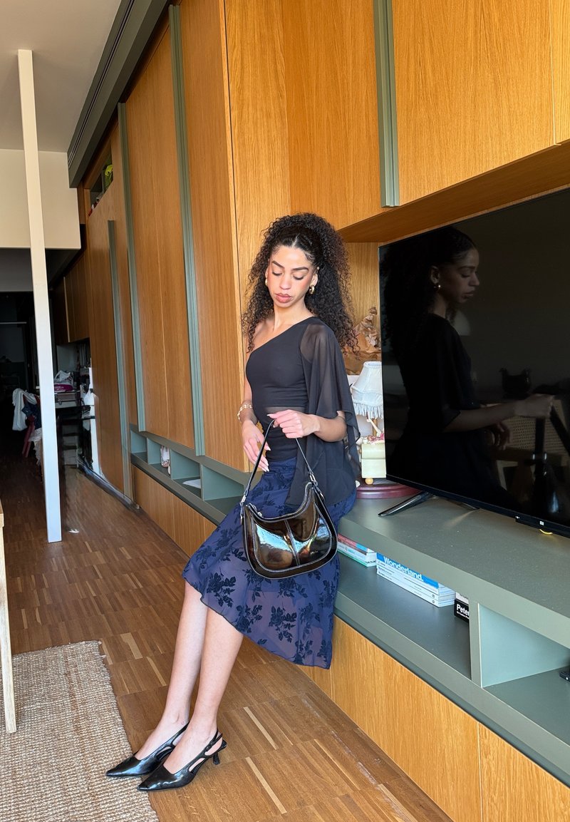Femme portant un haut noir asymétrique à une épaule et une jupe marine à fleurs, tenant un sac à main en cuir verni, assise sur un meuble vert à côté d'une télévision.