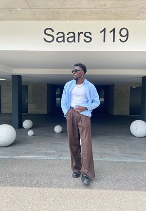 Hombre con gafas de sol y collar de cadena lleva pantalones marrones holgados, una camiseta sin mangas blanca y una camisa a rayas azules, de pie bajo un edificio con la etiqueta "Saars 119."