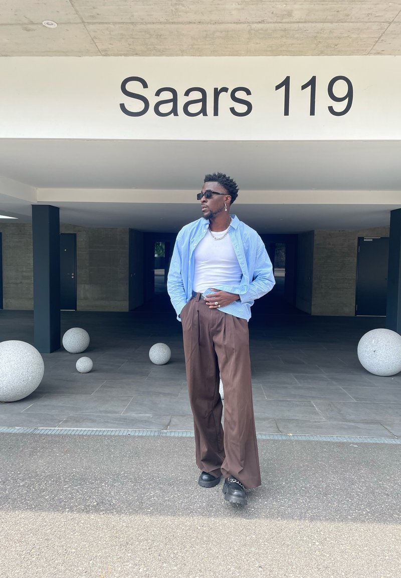 Homem com óculos de sol e colar de corrente usa calças largas castanhas, uma t-shirt branca e uma camisa de riscas azuis, parado debaixo de um edifício rotulado como "Saars 119".
