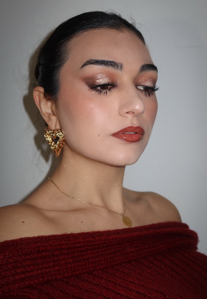 Le mannequin porte un haut bordeaux décolleté aux épaules, des boucles d'oreilles en forme de cœur doré et un collier en or délicat. Le maquillage met en valeur des tons bronze et des lèvres brillantes.