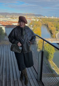 Smeđa kožna jakna, zelena marama oko vrata, crne široke hlače, smešak bereta i krznena torbica. Drvena terasa i pogled na reku u pozadini.