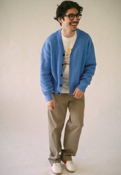 Cárdigan de punto azul claro con botones en la parte delantera, llevado sobre una camiseta gráfica blanca. Combinado con pantalones beige y zapatos blancos con acentos rojos.