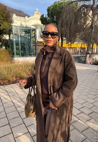 Femme portant un long manteau marron foncé et des lunettes de soleil, debout sur un chemin pavé baigné de soleil, à côté d'arbres et d'un ascenseur en verre.