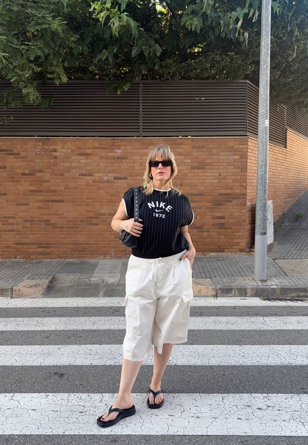 Melnbaltas svītrainas īsām piedurknēm Nike krekls, baltas kravas šortus, melni čībiņas un mazs melns soma pie ķieģeļu sienas.