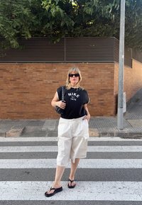 Melnbaltas svītrainas īsām piedurknēm Nike krekls, baltas kravas šortus, melni čībiņas un mazs melns soma pie ķieģeļu sienas.