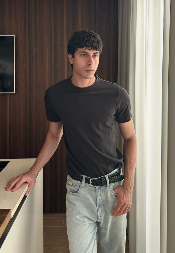 Jeune homme aux cheveux bouclés et foncés, portant un t-shirt noir et un jean clair, debout à l'intérieur près d'un comptoir et de rideaux blancs.