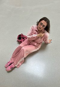 Rózsaszín selyem tréningruha fehér csíkokkal, illő rózsaszín cipővel és rózsaszín táskával. Ülő személy rövid, hullámos hajjal és napszemüvegben.