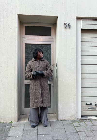 Hombre vestido con un abrigo largo marrón, guantes negros y pantalones grises, de pie frente a una puerta gris con el número 54 en una acera de la ciudad.