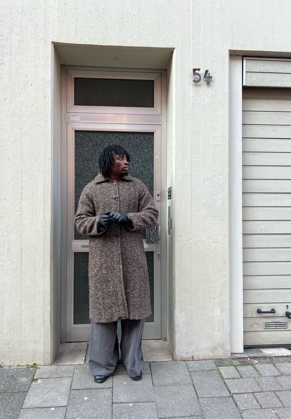 Egy férfi, hosszú barna kabátot, fekete kesztyűt és szürke nadrágot viselve, a városi járdán, egy 54-es számú szürke ajtó előtt áll.