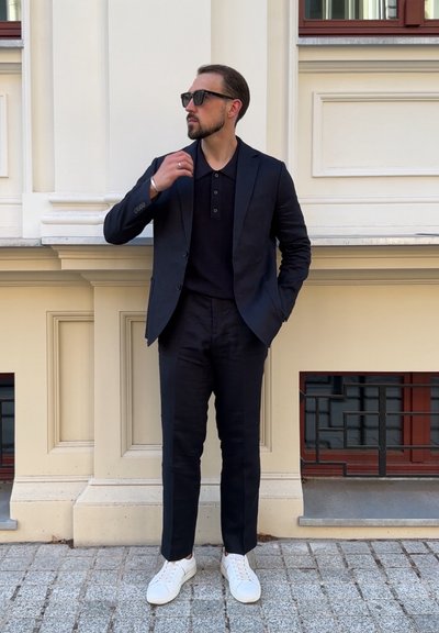 Traje azul marino con una camiseta polo negra, zapatillas blancas y gafas de sol. Ajuste slim y diseño estructurado, frente a una pared beige.