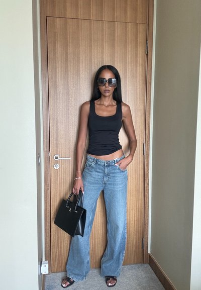 Top negro ajustado, jeans de tiro alto de pierna ancha en azul claro, bolso negro, gafas de sol negras de gran tamaño, de pie contra una puerta de madera.