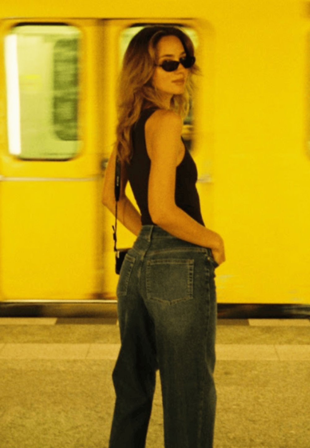 Hochgeschnittene blaue Jeans, figurbetontes schwarzes Tanktop, gelocktes Haar, Sonnenbrille, mit einer gelben U-Bahn im Hintergrund.