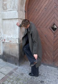 Personne en manteau gris long ajustant ou vérifiant l'ourlet d'un pantalon noir, debout à côté d'une grande porte en bois et d'un mur en pierre.