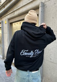 Zwarte hoodie met witte grafische tekst "Eternally Blessed" op de achterkant; gecombineerd met lichtblauwe spijkerbroek. De stof lijkt zacht en dik.