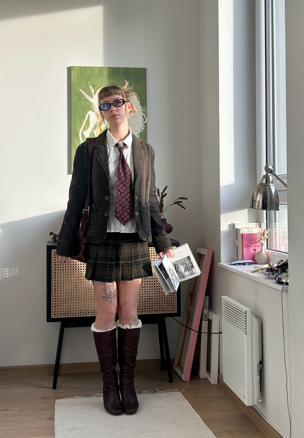 Brun kordfløyelsblazer over hvit skjorte og rødt mønstret slips, grønt rutet skjørt, brun knelange støvler, holder bøker, med synlige tatoveringer.