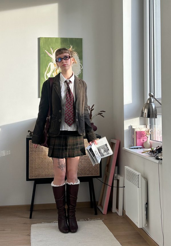 Sacou maro din catifea peste o cămașă albă și cravată roșie cu model, fustă verde în carouri, cizme maro până la genunchi, ținând cărți, cu tatuaje vizibile.