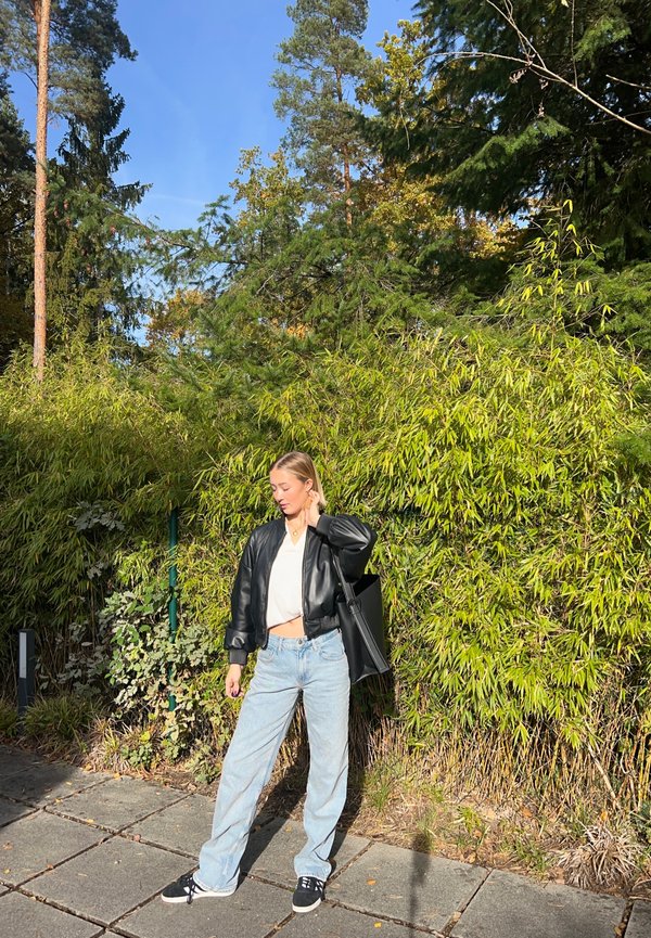 Chaqueta de cuero negra corta, camiseta blanca, jeans holgados azul claro y zapatillas negras; de pie frente a un bambú verde y árboles.