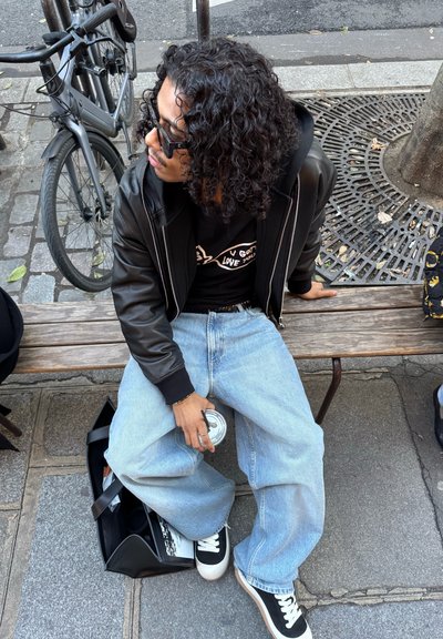 Persona con cabello rizado y gafas sentada en un banco de madera, sosteniendo una bebida, vistiendo chaqueta negra, camiseta gráfica, jeans claros y zapatillas negras.