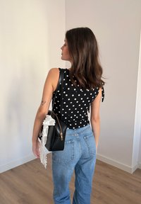 Femme aux cheveux bruns mi-longs portant un haut noir à pois et un jean bleu, tenant un sac à main noir avec un accessoire en dentelle blanche.