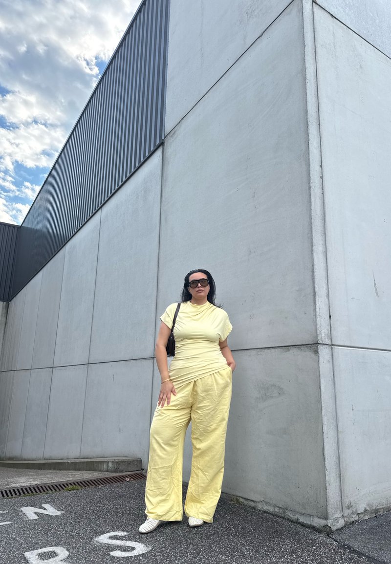 Woman wearing yellow outfit and sunglasses stands with hand in pocket against large grey concrete wall under partly cloudy sky.