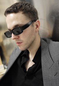 Lunettes de soleil rectangulaires noires avec une monture épaisse, portées avec une chemise noire et une veste de costume grise à texture. Fond neutre visible.